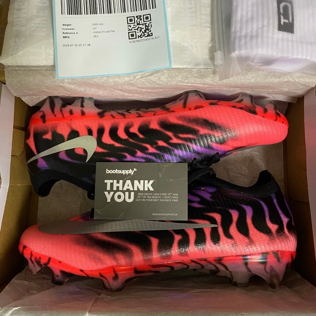 A pair of Nike soccer cleats with a vibrant neon orange, purple, and black wavy pattern, displayed inside their cardboard box alongside a white branded shoe bag, a shipping label, and a boot supply thank you card.