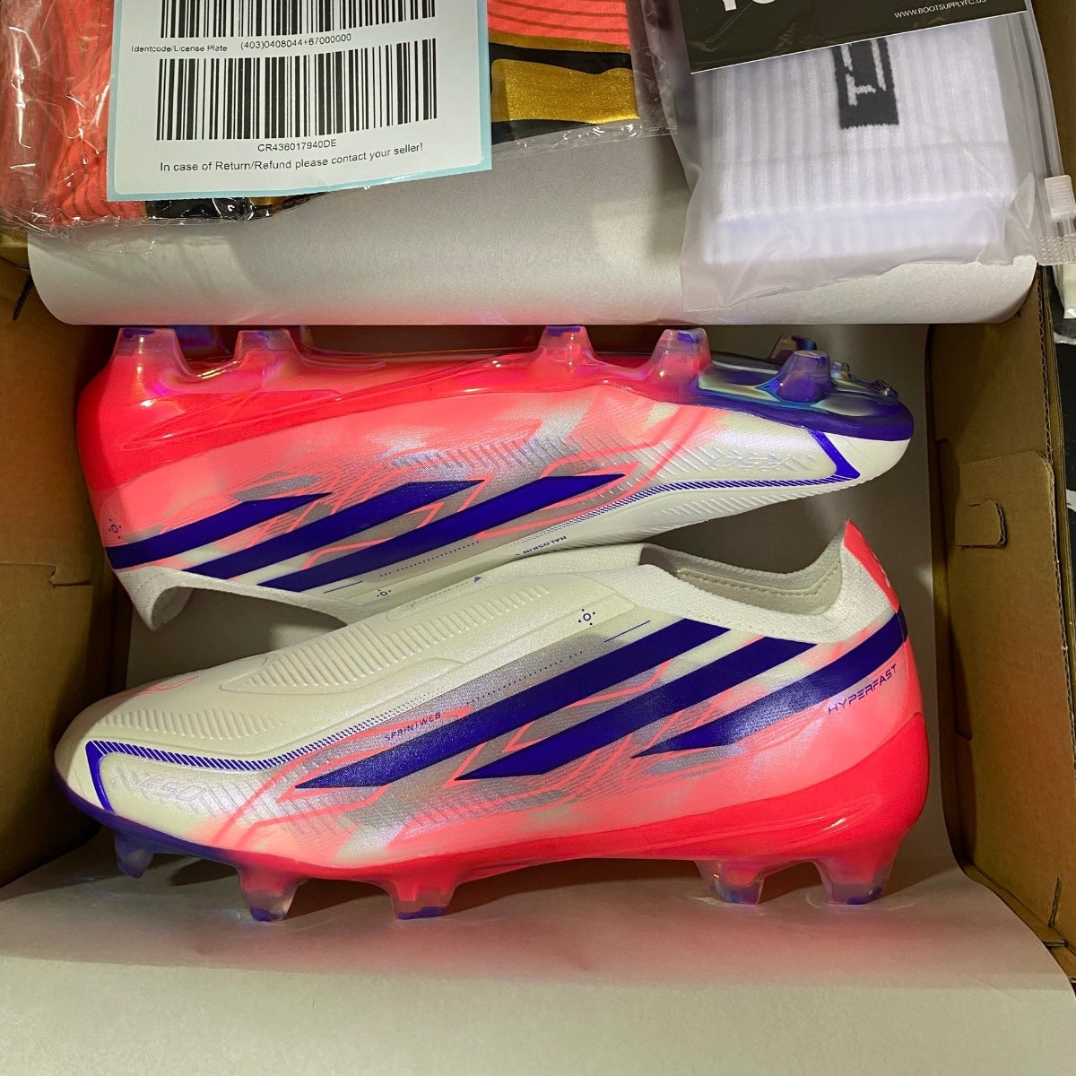 A pair of soccer cleats in a cardboard box, with white socks and a package of shin guards also visible. The cleats are white with bright pink/coral overlays and blue stripes.